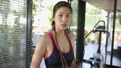 Serious-asian-woman-with-skipping-rope-on-neck-looking-at-camera-in-sunny-home-gym,-slow-motion