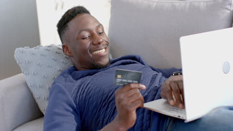 Feliz-Hombre-Afroamericano-Acostado-Y-Usando-Laptop-Y-Tarjeta-De-Crédito-En-Una-Soleada-Sala-De-Estar,-Cámara-Lenta