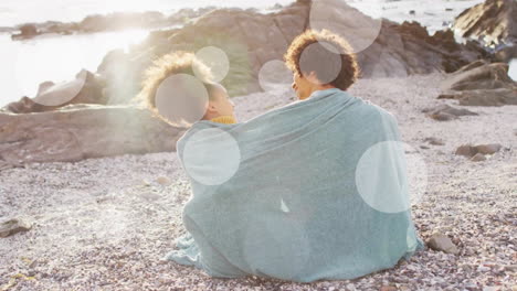 Animation-of-light-spots-over-biracial-couple-covered-in-blanket-embracing-on-beach