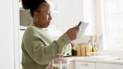 Happy-african-american-senior-woman-drinking-coffee,-using-tablet-in-sunny-kitchen,-slow-motion