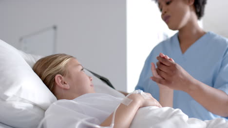 Diverse-female-doctor-and-girl-patient-lying-on-bed-holding-hands-in-hospital,-slow-motion