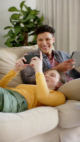 Vertical-video-of-happy-diverse-couple-sitting-on-sofa-using-tablet-and-smartphone,-in-slow-motion