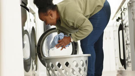 Glückliche-Afroamerikanische-Seniorin,-Die-Wäsche-In-Die-Waschmaschine-In-Einer-Sonnigen-Küche-Legt,-Zeitlupe
