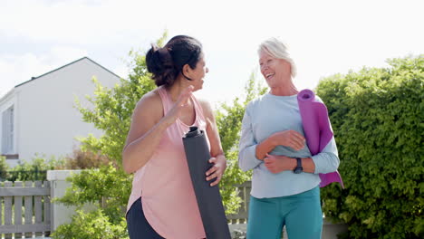 Two-happy-diverse-senior-women-with-exercise-mats,-embracing-in-garden,-slow-motion,-copy-space