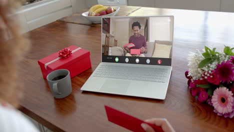 Caucasian-woman-holding-red-envelope-using-laptop-with-african-american-man-with-gift-on-screen