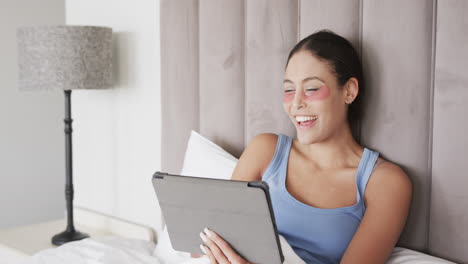Happy-biracial-woman-with-under-eye-patches-sitting-on-bed-using-tablet,-slow-motion