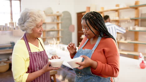 Happy-diverse-female-potters-holding-bowls-and-discussing-in-pottery-studio,-slow-motion