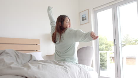 Happy-plus-size-biracial-woman-sitting-on-bed-stretching-in-the-morning,-copy-space,-slow-motion
