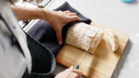 Biracial-woman-wearing-apron-and-cutting-fresh-bread-in-kitchen,-slow-motion