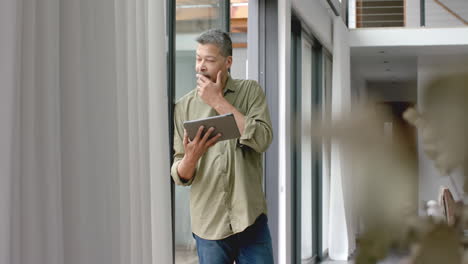 Thoughtful-senior-biracial-man-standing-by-window-using-tablet-at-home,-copy-space,-slow-motion