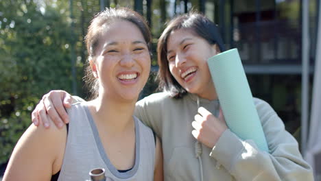Happy-asian-female-friends-with-exercise-mats-and-water-bottle-laughing-on-terrace,-slow-motion