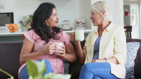 Happy-diverse-senior-women-with-coffee,-discussing-on-sofa-in-sunny-living-room,-slow-motion