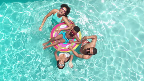 Aerial-of-african-american-parents,-son-and-daughter-on-inflatable-in-sunny-pool-waving,-copy-space