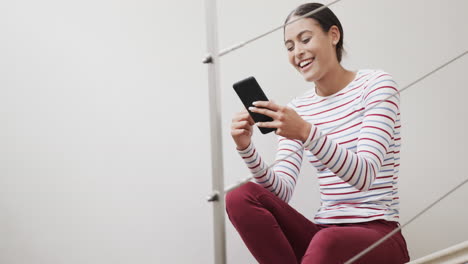Happy-biracial-woman-sitting-on-stairs-using-smartphone,-slow-motion