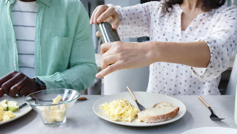 Feliz-Pareja-Birracial-Sazonando-Y-Desayunando-Huevos-Revueltos-En-La-Cocina,-Cámara-Lenta