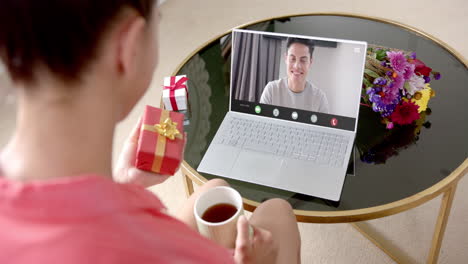 Caucasian-woman-holding-red-gift-using-laptop-with-caucasian-man-on-screen
