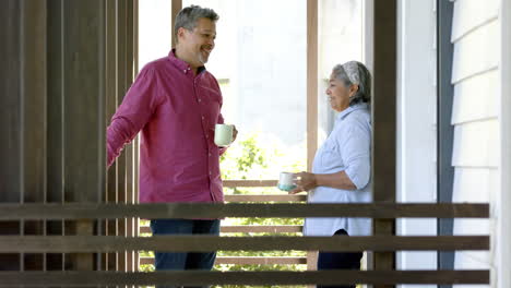 Happy-biracial-senior-couple-standing-in-window,-talking-and-drinking-tea