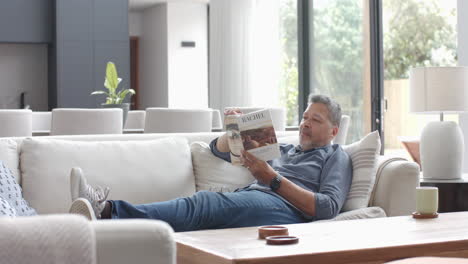 Focused-senior-biracial-man-relaxing-on-couch-reading-book-in-living-room,-slow-motion
