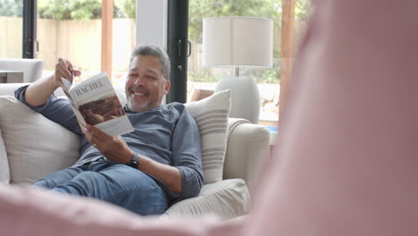 Happy-senior-biracial-man-relaxing-on-couch-reading-book-in-living-room,-copy-space,-slow-motion
