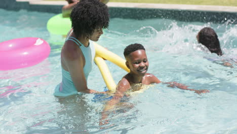 Happy-african-american-mother-teaching-son-to-swim-with-noodle-float-in-sunny-pool,-slow-motion