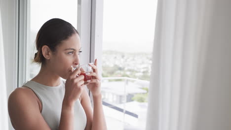 Thoughtful-biracial-woman-looking-through-window-drinking-tea-at-home,-copy-space,-slow-motion