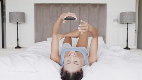 Happy-biracial-woman-lying-on-bed-using-smartphone,-slow-motion