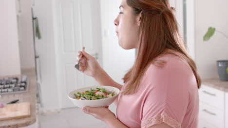 Happy-plus-size-biracial-woman-eating-vegetable-salad-in-kitchen,-slow-motion
