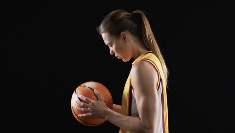 Focused-young-Caucasian-woman-holds-a-basketball-in-a-dark-setting-on-a-black-background,-conveying-