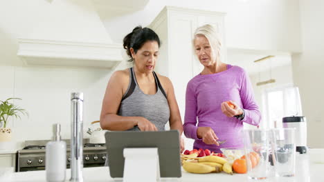 Dos-Mujeres-Mayores-Felices-Y-Diversas-Preparando-Frutas-Usando-Tableta-En-La-Cocina,-Cámara-Lenta