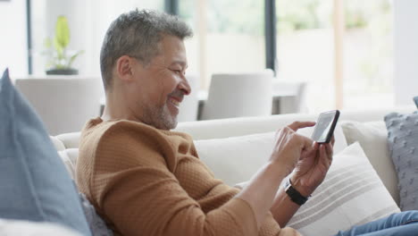 Glücklicher-älterer-Mann-Mit-Gemischter-Abstammung,-Der-Sein-Smartphone-Auf-Der-Couch-Benutzt,-Im-Sonnigen-Wohnzimmer-Lacht,-Zeitlupe