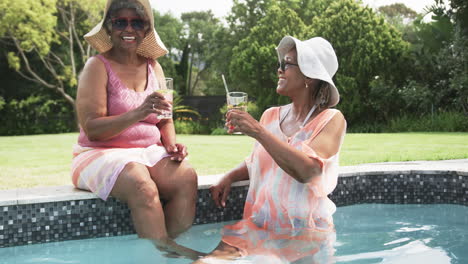 Glückliche-ältere-Afroamerikanische-Freundinnen-In-Sonnenhüten,-Die-Mit-Getränken-Im-Pool-Sitzen,-Zeitlupe