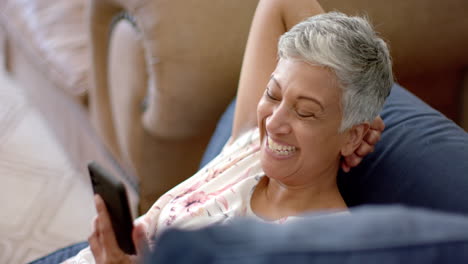Glückliche-ältere-Frau-Mit-Gemischter-Abstammung,-Die-Auf-Der-Couch-Liegt-Und-Zu-Hause-Mit-Dem-Smartphone-Spricht,-Zeitlupe