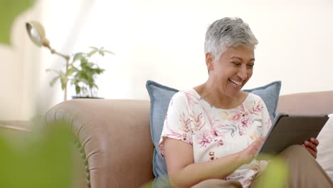 Glückliche-ältere-Frau-Mit-Gemischter-Abstammung,-Die-Lachend-Auf-Der-Couch-Sitzt-Und-Zu-Hause-Ein-Tablet-Benutzt,-Zeitlupe