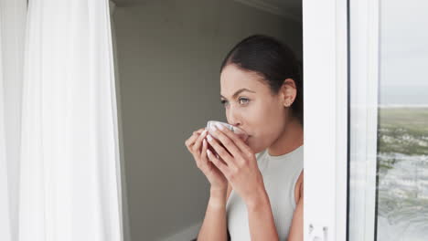 Thoughtful-biracial-woman-looking-through-window-drinking-tea-at-home,-slow-motion