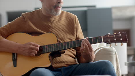 Hombre-Birracial-Senior-Enfocado-Sentado-En-El-Sofá-Tocando-La-Guitarra-En-Casa,-Cámara-Lenta