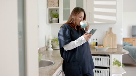 Happy-plus-size-biracial-female-doctor-with-coffee-using-smartphone-at-home,-copy-space,-slow-motion