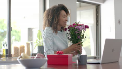Glückliche-Biracial-Frau-Mit-Geschenk-Und-Blumen,-Die-Zu-Hause-Einen-Videoanruf-Auf-Dem-Laptop-Führt,-Zeitlupe