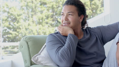Happy,-thoughtful-biracial-man-sitting-in-sunny-living-room-enjoying-view,-copy-space,-slow-motion