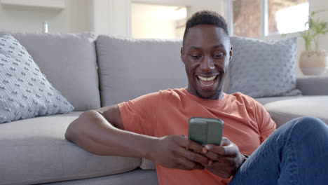 Hombre-Afroamericano-Feliz-Teniendo-Videollamada-En-Un-Teléfono-Inteligente-En-Una-Sala-De-Estar-Soleada,-Cámara-Lenta
