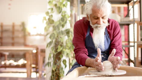 Focused-senior-biracial-potter-with-long-beard-using-potter's-wheel-in-pottery-studio,-slow-motion