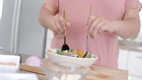 Midsection-of-plus-size-biracial-woman-tossing-bowl-of-feta-salad-in-kitchen,-slow-motion