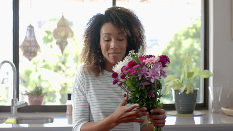 Happy-biracial-woman-with-gift-and-flowers-having-video-call-in-kitchen,-slow-motion