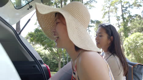 Happy-asian-female-friends-putting-suitcases-into-car-trunk-and-laughing-on-sunny-day,-slow-motion