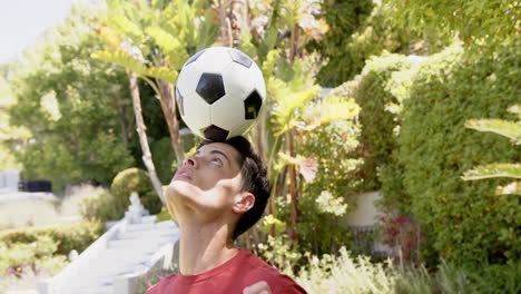 Hombre-Birracial-Enfocado-Practicando-Fútbol-De-Equilibrio-En-La-Cabeza-En-Un-Jardín-Soleado,-Cámara-Lenta