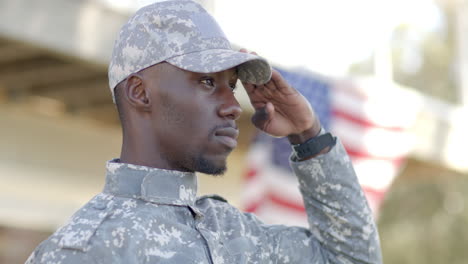 Ernster-Afroamerikanischer-Soldat-Salutiert-Mit-Flagge-Im-Hintergrund-An-Einem-Sonnigen-Tag,-Zeitlupe