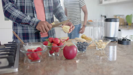 Happy-diverse-gay-male-couple-preparing-fruit-for-smoothie-in-kitchen,-copy-space,-slow-motion