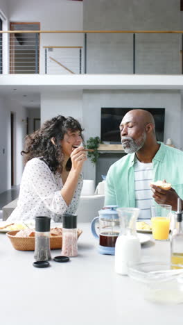 Vertical-video-of-happy-diverse-mature-couple-eating-breakfast-together-at-home,-slow-motion