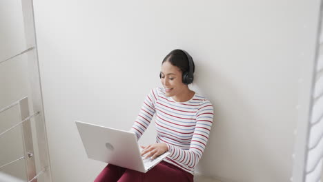 Happy-biracial-woman-sitting-on-stairs-using-headphones-and-laptop,-slow-motion