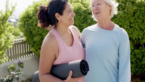 Two-happy-diverse-senior-women-with-exercise-mats,-embracing-and-smiling-in-garden,-slow-motion