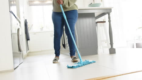Happy-african-american-senior-woman-cleaning-floor-and-smiling-in-sunny-kitchen,-slow-motion